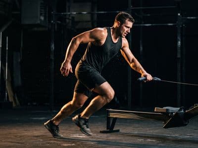 An athlete stepping off a rowing machine straight into a run, mid-transition, one hand still on the handle, motion in the legs