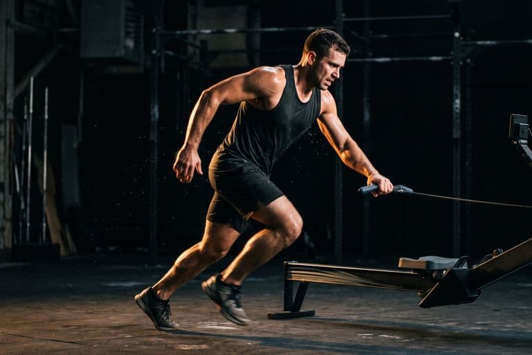 An athlete stepping off a rowing machine straight into a run, mid-transition, one hand still on the handle, motion in the legs