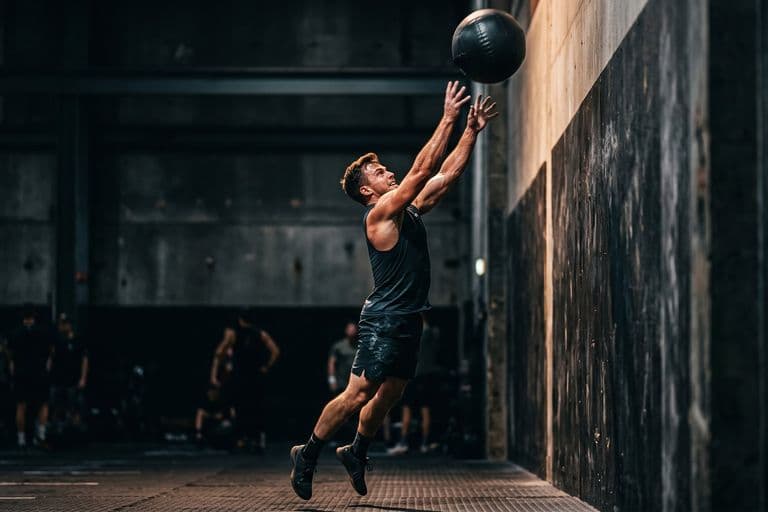An athlete mid-wall-ball throw with a heavy medicine ball at full extension overhead, hard side light, dark hall behind