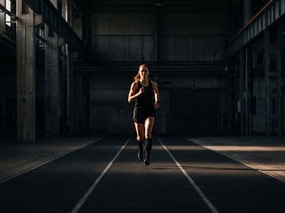 An athlete running alone on a marked indoor course in a dark hall, seen head-on at distance, deliberately controlled rather than sprinting