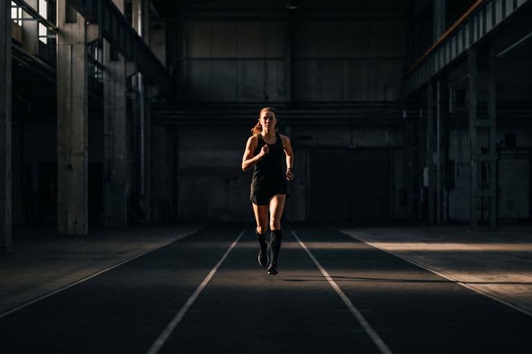 An athlete running alone on a marked indoor course in a dark hall, seen head-on at distance, deliberately controlled rather than sprinting