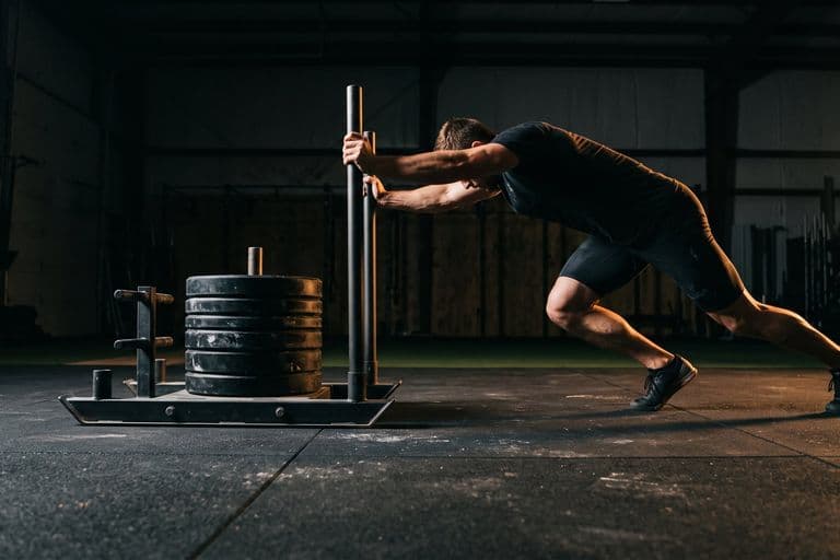A tight low ground-level view of a loaded sled and an athlete's driving legs and locked arms, body angle almost horizontal, matting texture sharp