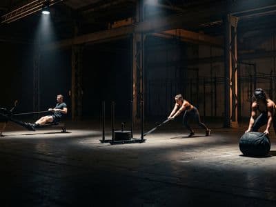 A wide view of a dark indoor hall with several athletes working at spaced unbranded stations — a rower, a sled, a sandbag — pools of hard light on each